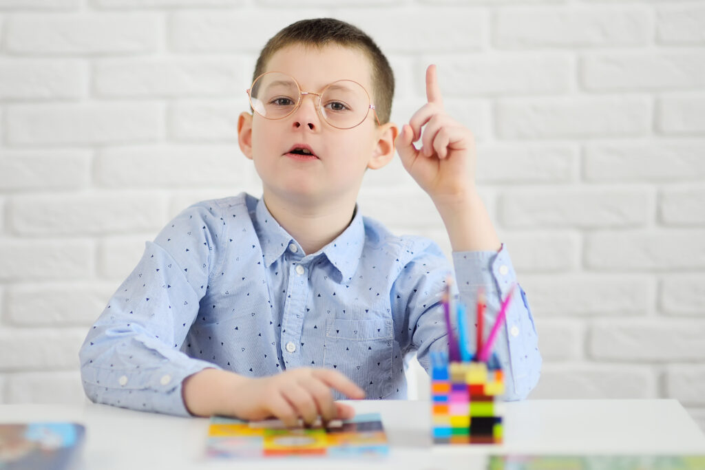 A Schoolboy Child With Glasses Sitting At A White Table Shows A Gesture An Index Finger Up.